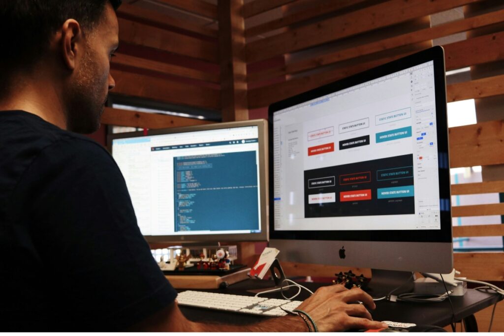 Programista pracujący przy dwóch monitorach, co symbolizuje proces rozwoju strony internetowej – od projektowania interfejsu (UX/UI) po kodowanie.